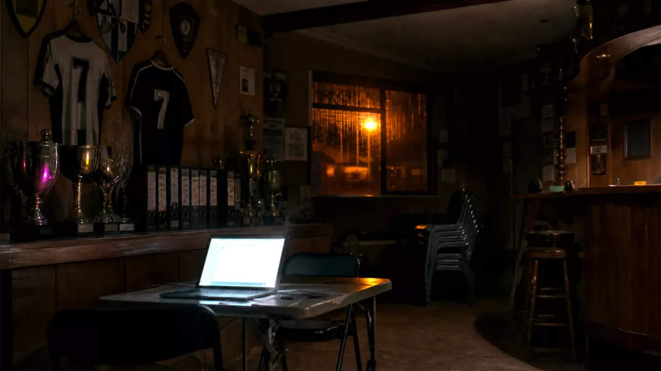 Local de club la nuit, trophées et classeurs dans l’ombre, un vieil ordinateur portable ouvert sur une table pliante, seule lumière de la pièce.