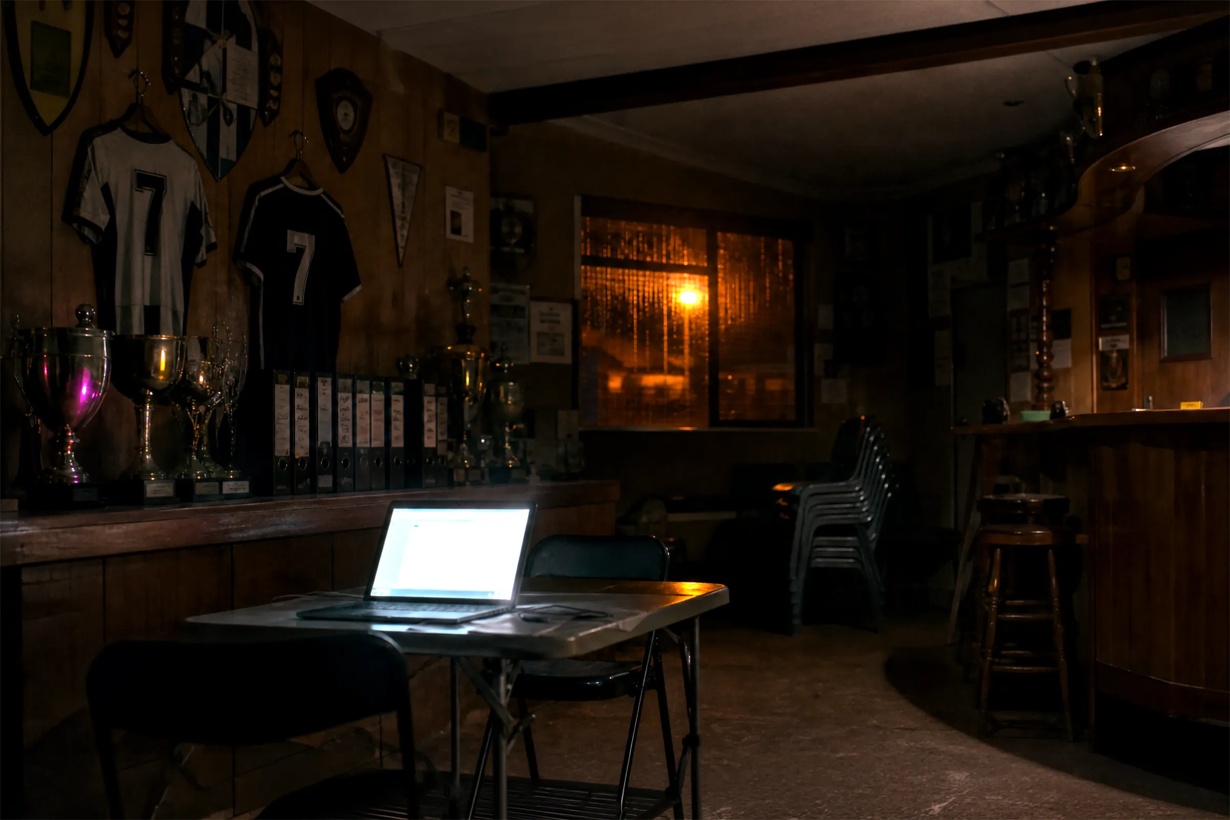 Local de club la nuit, trophées et classeurs dans l’ombre, un vieil ordinateur portable ouvert sur une table pliante, seule lumière de la pièce.